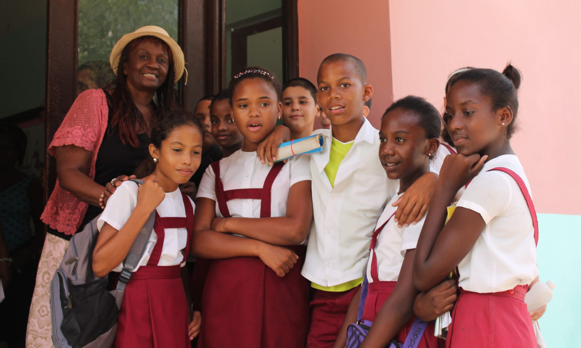A woman in a straw hat with a group of students in school uniforms posing together outside a school doorway during a Simunye outreach trip.