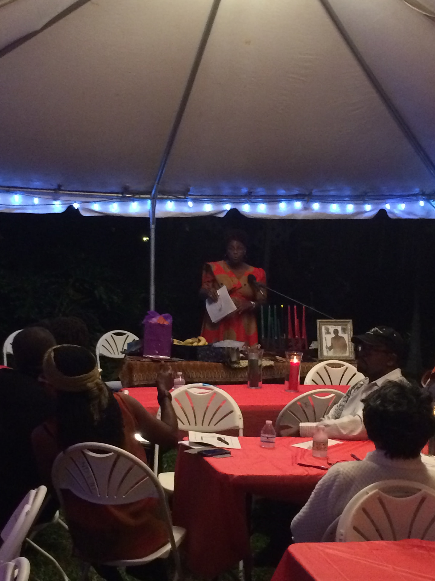 A woman in a red-and-orange dress speaking to guests from a head table with a kinara under a white tent decorated with lights during a Simunye Kwanzaa celebration.