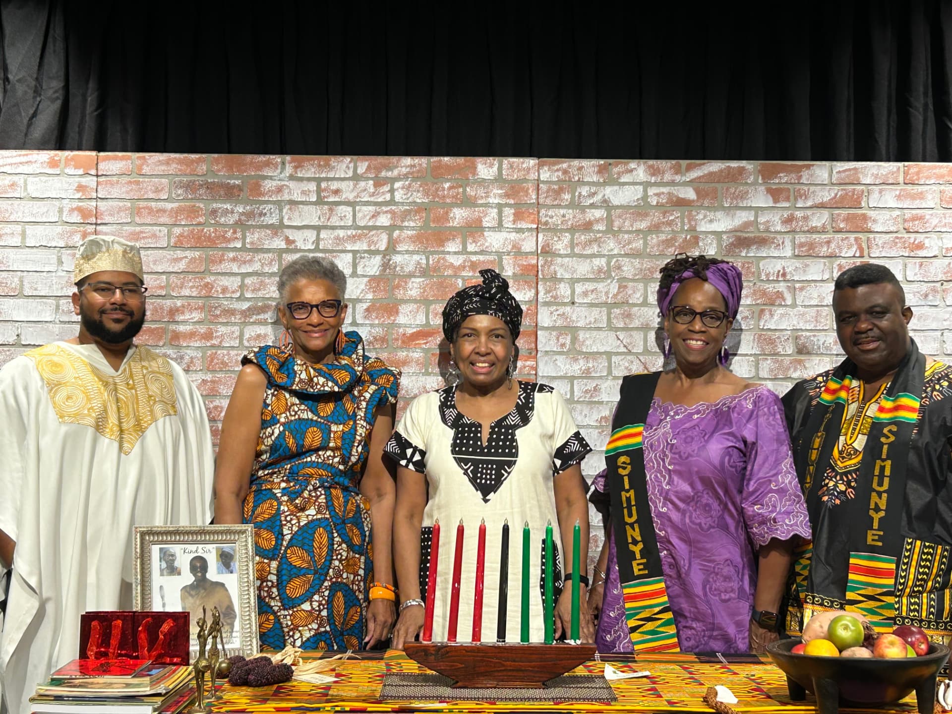 Five Simunye members wearing SIMUNYE sashes standing behind a Kente-cloth-covered table holding a kinara, a framed photo, fruit, and books at a Kwanzaa celebration.